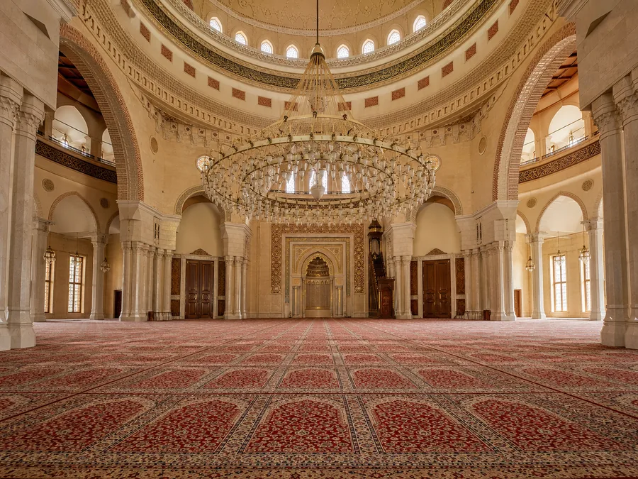 قاعة الصلاة المفروشة بالسجاد في جامع أحمد الفاتح تحت قبته، مع ثريا ضخمة مزخرفة وأعمدة رخامية